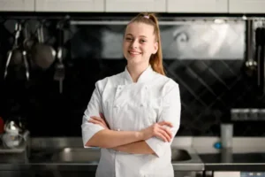 A professional female chef in a white uniform and hat, carefully plating a gourmet dish in a modern kitchen