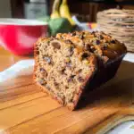 A slice of moist brown butter banana bread filled with chocolate chips on a wooden board