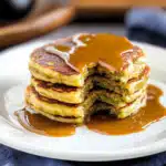 Stack of zucchini banana pancakes on a white plate, topped with syrup and a bite taken out.