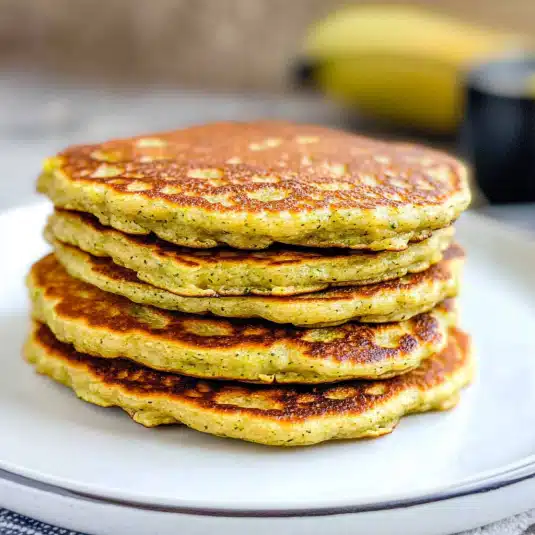 Stack of golden zucchini banana pancakes on a white plate, showing crisp edges and soft centers.