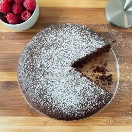 Whole almond flour chocolate cake with one slice missing, dusted with powdered sugar and served with fresh raspberries on a wooden board