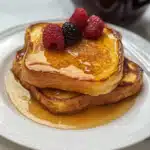 Golden brioche French toast stacked with maple syrup and fresh berries on a white plate