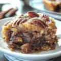 Close-up of a gooey pecan pie dump cake slice topped with toasted pecans, served on a white plate with a spoon beside it.