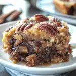 Close-up of a gooey pecan pie dump cake slice topped with toasted pecans, served on a white plate with a spoon beside it.